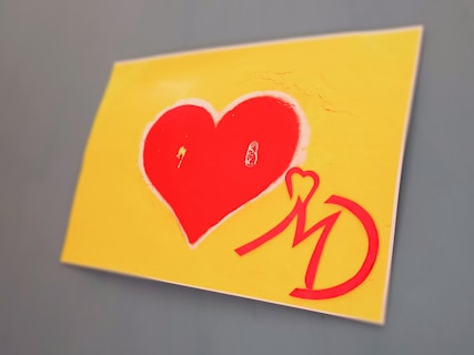 A piece of art featuring a large red heart against a yellow background. Below the heart, there is a stylized red 'MD' monogram with heart motifs incorporated into the letters, all set against a flat surface.