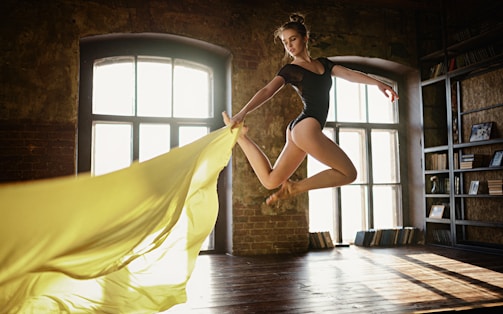 A dancer in a black leotard is captured in mid-air leap inside a spacious studio with large windows. The room is warmly lit, highlighting her graceful form and the flowing yellow fabric she holds, creating a dynamic sense of movement. The surroundings include exposed brick walls and shelves filled with books.