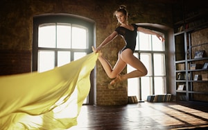 A dancer in a black leotard is captured in mid-air leap inside a spacious studio with large windows. The room is warmly lit, highlighting her graceful form and the flowing yellow fabric she holds, creating a dynamic sense of movement. The surroundings include exposed brick walls and shelves filled with books.