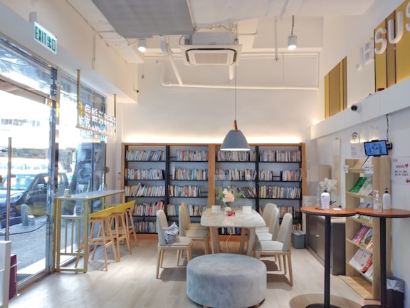 A cozy indoor space with a modern, minimalist design. Bookshelves filled with colorful books line the back wall. In the center, there is a round table surrounded by chairs and topped with a vase of flowers. To the left, a bar-style seating area features stools by a window. The lighting is soft and warm, creating a comfortable atmosphere.