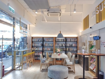 A cozy indoor space with a modern, minimalist design. Bookshelves filled with colorful books line the back wall. In the center, there is a round table surrounded by chairs and topped with a vase of flowers. To the left, a bar-style seating area features stools by a window. The lighting is soft and warm, creating a comfortable atmosphere.