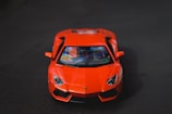 A bright red sports car is prominently displayed against a dark background, with a focus on its sleek and aerodynamic front design. The car's glossy surface reflects lighting, adding a dynamic range to its appearance.