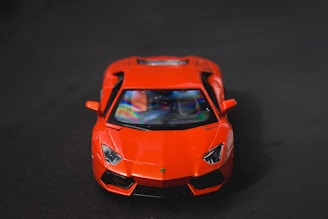 A bright red sports car is prominently displayed against a dark background, with a focus on its sleek and aerodynamic front design. The car's glossy surface reflects lighting, adding a dynamic range to its appearance.