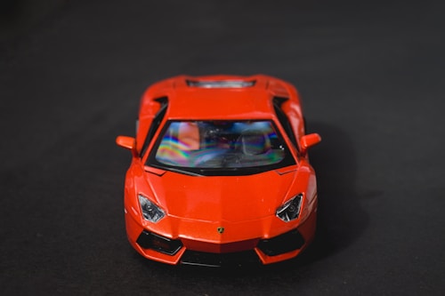 A bright red sports car is prominently displayed against a dark background, with a focus on its sleek and aerodynamic front design. The car's glossy surface reflects lighting, adding a dynamic range to its appearance.