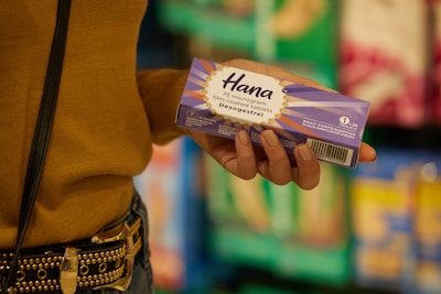 A person holds a purple box labeled 'Hana' which contains 75 microgram film-coated tablets of desogestrel, a daily contraceptive. The background features blurred shelves of various products, suggesting a store setting. The person's clothing includes a mustard-colored top and a studded belt.