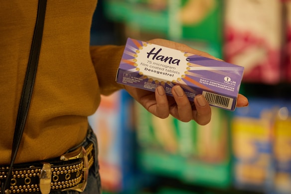 A person holds a purple box labeled 'Hana' which contains 75 microgram film-coated tablets of desogestrel, a daily contraceptive. The background features blurred shelves of various products, suggesting a store setting. The person's clothing includes a mustard-colored top and a studded belt.