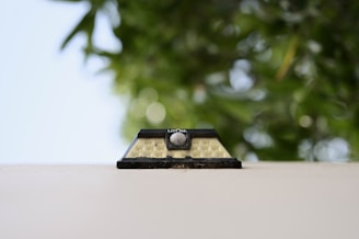 A small solar wall lamp is attached to an off-white surface, set against a blurred backdrop of lush green leaves. The design is simple with a rectangular shape and a central circular sensor.