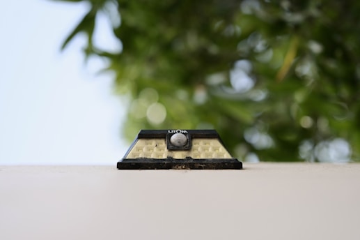 A small solar wall lamp is attached to an off-white surface, set against a blurred backdrop of lush green leaves. The design is simple with a rectangular shape and a central circular sensor.