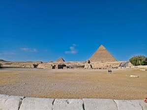 A wide shot of the Egyptian desert, featuring the Great Pyramids and the Sphinx under a clear blue sky. Several people are scattered across the foreground, exploring the ancient site. The arid landscape surrounds these iconic monuments, and the vastness of the scene conveys the historical significance and grandeur of the location.