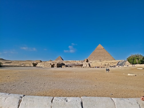 A wide shot of the Egyptian desert, featuring the Great Pyramids and the Sphinx under a clear blue sky. Several people are scattered across the foreground, exploring the ancient site. The arid landscape surrounds these iconic monuments, and the vastness of the scene conveys the historical significance and grandeur of the location.