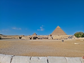 A wide shot of the Egyptian desert, featuring the Great Pyramids and the Sphinx under a clear blue sky. Several people are scattered across the foreground, exploring the ancient site. The arid landscape surrounds these iconic monuments, and the vastness of the scene conveys the historical significance and grandeur of the location.
