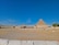 A wide shot of the Egyptian desert, featuring the Great Pyramids and the Sphinx under a clear blue sky. Several people are scattered across the foreground, exploring the ancient site. The arid landscape surrounds these iconic monuments, and the vastness of the scene conveys the historical significance and grandeur of the location.