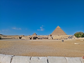 A wide shot of the Egyptian desert, featuring the Great Pyramids and the Sphinx under a clear blue sky. Several people are scattered across the foreground, exploring the ancient site. The arid landscape surrounds these iconic monuments, and the vastness of the scene conveys the historical significance and grandeur of the location.