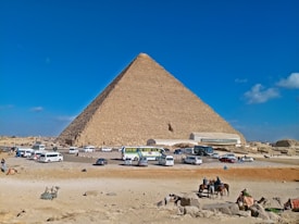 A large pyramid stands majestically against a bright blue sky, surrounded by a sandy desert landscape. Several cars and buses are parked nearby, and people on horseback are visible in the foreground. The scene includes tourists observing the historic site with a few camels resting on the ground.