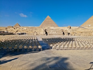 Rows of empty chairs are arranged on a stone platform in front of the iconic pyramids of Giza, with a clear blue sky above. The Sphinx is visible in the background, facing the seating arrangement.