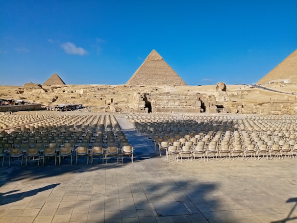 Rows of empty chairs are arranged on a stone platform in front of the iconic pyramids of Giza, with a clear blue sky above. The Sphinx is visible in the background, facing the seating arrangement.