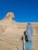 A person wearing a headscarf and jacket is looking at the Great Sphinx of Giza in a desert landscape. The sky is clear and blue, and there are several chains creating a barrier along the viewing area.