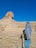 A person wearing a headscarf and jacket is looking at the Great Sphinx of Giza in a desert landscape. The sky is clear and blue, and there are several chains creating a barrier along the viewing area.