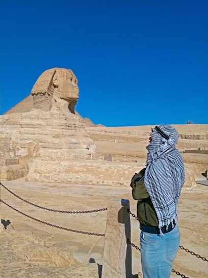 A person wearing a headscarf and jacket is looking at the Great Sphinx of Giza in a desert landscape. The sky is clear and blue, and there are several chains creating a barrier along the viewing area.