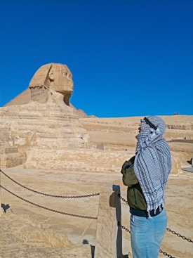 A person wearing a headscarf and jacket is looking at the Great Sphinx of Giza in a desert landscape. The sky is clear and blue, and there are several chains creating a barrier along the viewing area.