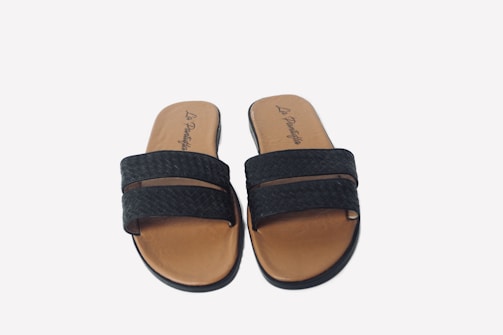 A pair of black woven sandals with brown soles, featuring two straps across the foot. The inner sole displays a logo with stylish script.