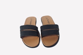 A pair of black woven sandals with brown soles, featuring two straps across the foot. The inner sole displays a logo with stylish script.
