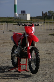 A red dirt bike with thick off-road tires is positioned on a sandy area. The background features barriers, a white and green cylindrical structure, and some distant buildings and greenery.