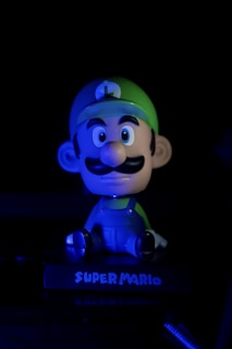 A cartoonish figurine with a large head, wearing a green cap and blue overalls, is illuminated with a moody blue light against a dark background. The base of the figurine reads 'Super Mario.'