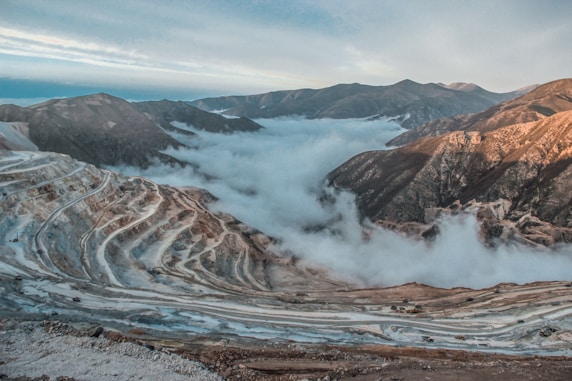 Terraced layers of an open-pit mine stretch across the rugged terrain, with swirling clouds nestled between the steep mountain ranges that frame the horizon. The rugged surface is marked by the distinct lines and curves of mining operations, contrasting with the smooth, ethereal mist that blankets the landscape, creating a dramatic interplay between industry and nature.