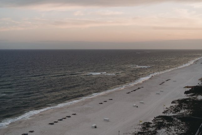 A serene beach at sunset with gentle waves washing ashore. The sand is dotted with evenly spaced rows of dark beach loungers. The horizon is painted with soft hues of orange and pink. There are empty white cabanas scattered along the sand, and a few patches of vegetation are visible in the foreground.