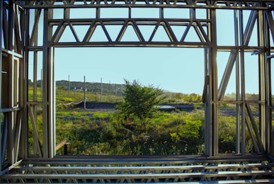 A view through a metal framework structure revealing a natural landscape. The foreground is dominated by the geometric lines of the framework, which contrasts with the softer, more organic forms of the grass and trees outside. Beyond the structure, there is lush greenery, including a prominent tree in the midground and a distant view of hills under a clear blue sky.
