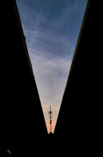 A silhouette of a communication tower is framed between two converging dark structures, set against a backdrop of a blue sky with soft, wispy clouds and a warm glow near the horizon.