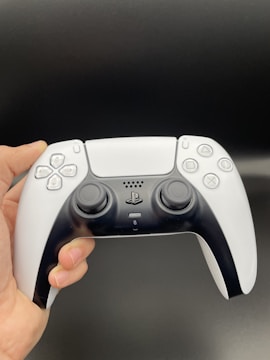 A person holds a modern gaming controller with a sleek design. The controller features two analog sticks, a directional pad, and various iconic buttons. The color scheme combines white and black with a smooth finish.
