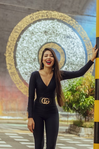 A woman with long hair is smiling, wearing a black outfit with a prominent belt. She is standing outdoors in front of a large decorative circular mural. The mural features concentric patterns in white and gold against a gray background. There are plants to the side and a pole with yellow and black stripes near her.