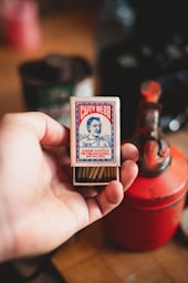 A hand holding an open matchbox with a vintage design. The box has a label featuring a portrait and the name 'Capt Webb', along with the text about swimming across the English Channel. In the background, there's a red metal object that resembles an old fuel can and a blurred backdrop of various items.
