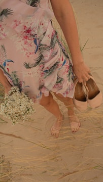 A person standing barefoot on a sandy surface, wearing a floral dress and holding a pair of shoes in one hand, along with a small bouquet of white flowers in the other hand. The setting includes some sparse grass around the sandy area.