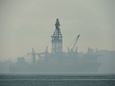 An industrial structure, likely an oil rig, sits in the middle of a body of water, partially obscured by heavy fog or mist. The silhouette reveals cranes and intricate scaffolding against a muted, misty backdrop. The water appears calm, with slight ripples visible in the foreground.