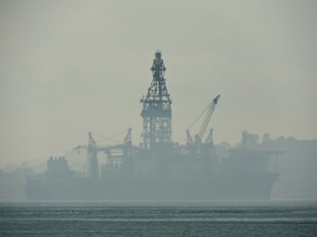 An industrial structure, likely an oil rig, sits in the middle of a body of water, partially obscured by heavy fog or mist. The silhouette reveals cranes and intricate scaffolding against a muted, misty backdrop. The water appears calm, with slight ripples visible in the foreground.
