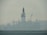 An industrial structure, likely an oil rig, sits in the middle of a body of water, partially obscured by heavy fog or mist. The silhouette reveals cranes and intricate scaffolding against a muted, misty backdrop. The water appears calm, with slight ripples visible in the foreground.