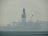 An industrial structure, likely an oil rig, sits in the middle of a body of water, partially obscured by heavy fog or mist. The silhouette reveals cranes and intricate scaffolding against a muted, misty backdrop. The water appears calm, with slight ripples visible in the foreground.