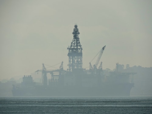 An industrial structure, likely an oil rig, sits in the middle of a body of water, partially obscured by heavy fog or mist. The silhouette reveals cranes and intricate scaffolding against a muted, misty backdrop. The water appears calm, with slight ripples visible in the foreground.