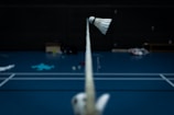 A badminton shuttlecock is balancing on a net in a dimly lit indoor court. The court has a blue floor with white lines marking the boundaries. Various items and sports equipment are scattered in the background, and the overall atmosphere is quiet and focused.