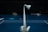 A badminton shuttlecock is balancing on a net in a dimly lit indoor court. The court has a blue floor with white lines marking the boundaries. Various items and sports equipment are scattered in the background, and the overall atmosphere is quiet and focused.