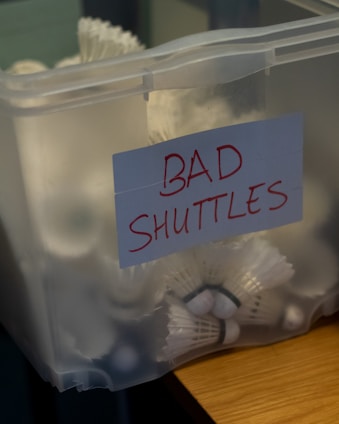 A transparent plastic container is filled with badminton shuttlecocks. A paper label on the container reads 'BAD SHUTTLES' in red marker, suggesting these shuttlecocks may be deemed unsuitable for use.