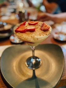 A glass filled with a creamy dessert topped with sliced strawberries and a sprinkle of cinnamon or cocoa. The dessert is elegantly presented on a large, dark-colored plate. The background is softly blurred, suggesting a warm and inviting dining environment.