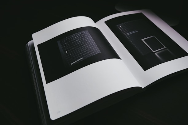 An open book displaying two dark, abstract images on its pages. The lighting is dim, emphasizing the minimalistic and modern design of the inside pages. The images feature geometric patterns and minimal text.
