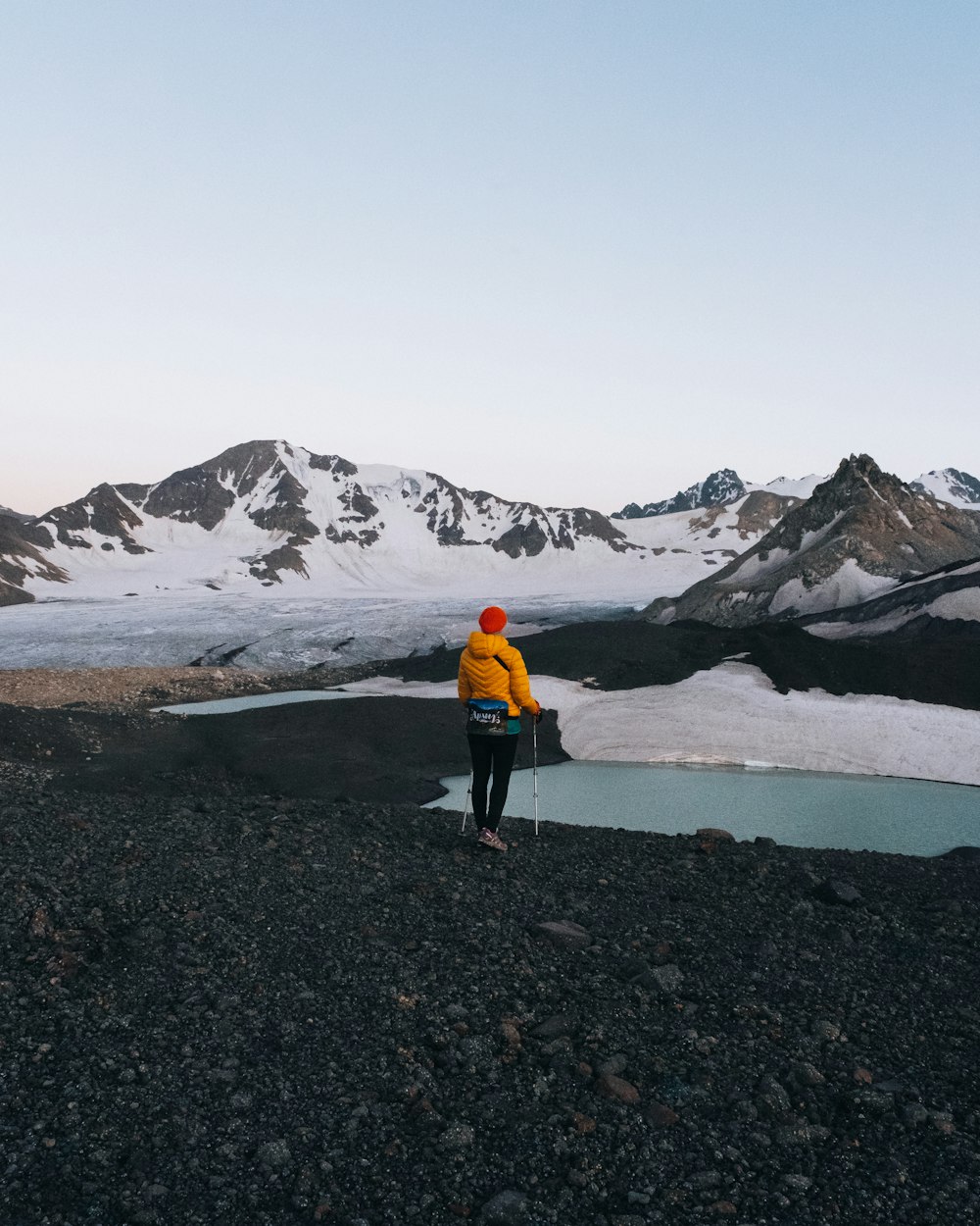 person in yellow jacket and black pants walking on gray sand near snow covered mountain during