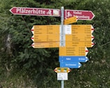 Numerous directional signs are clustered on a pole, providing information about hiking and cycling routes. The signs are colored in pink, yellow, and blue, and include distance and estimated time durations to various destinations such as Pfälzerhütte, Vaduz, and Triesenberg. These signs are set against a backdrop of lush greenery.