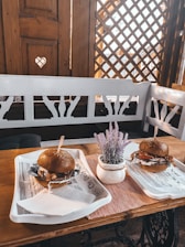 A cozy restaurant setting featuring a wooden table and white bench seating. On the table, there are two trays displaying burgers with sesame seed buns, filled with a variety of ingredients like lettuce and sauce. A small pot with lavender is placed between the trays. The wooden accents and decorative lattice on the walls give a warm, rustic feel.