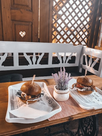 A cozy restaurant setting featuring a wooden table and white bench seating. On the table, there are two trays displaying burgers with sesame seed buns, filled with a variety of ingredients like lettuce and sauce. A small pot with lavender is placed between the trays. The wooden accents and decorative lattice on the walls give a warm, rustic feel.